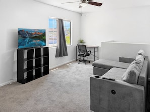 A modern living room with a flat-screen TV, a grey sofa, a black office chair, and a ceiling fan. - Quiet Casa Grande 4BR Gem | Snowbird Winter Base (Casa Grande)