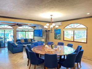 A spacious living area with a round wooden table surrounded by chairs, a sofa set, and a large window offering a view of the outdoors. - Jungle Pool Villa - 5BR Gated Playacar Hideaway (Playa del Carmen)