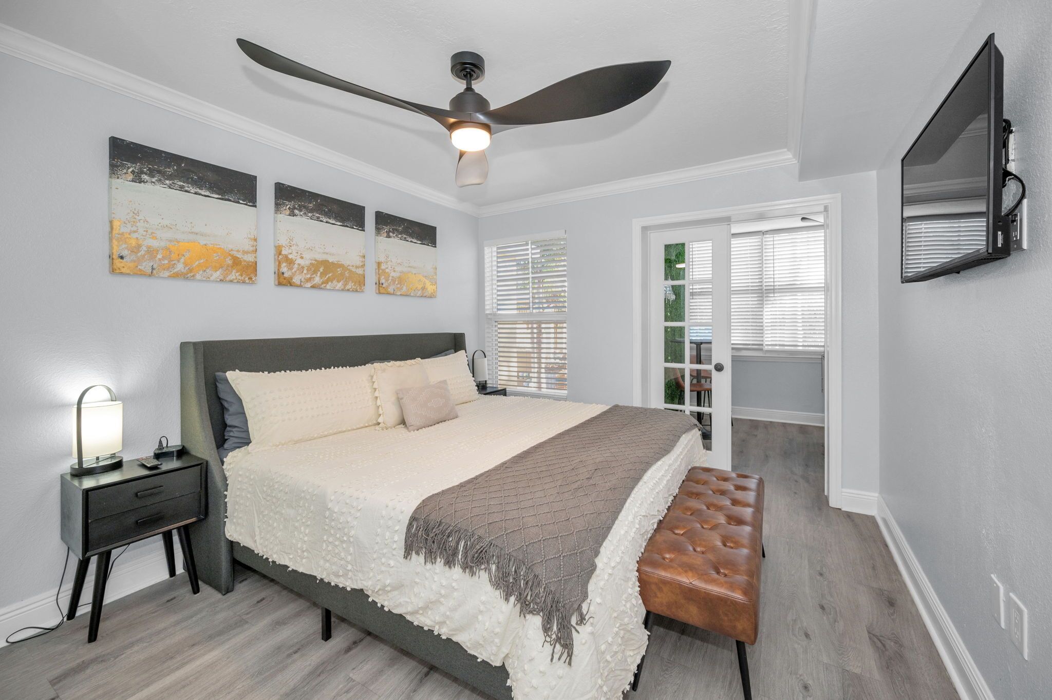 A bedroom with a bed, nightstand, lamp, ceiling fan, artwork, and a TV mounted on the wall.