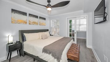 A bedroom with a bed, nightstand, lamp, ceiling fan, artwork, and a TV mounted on the wall.