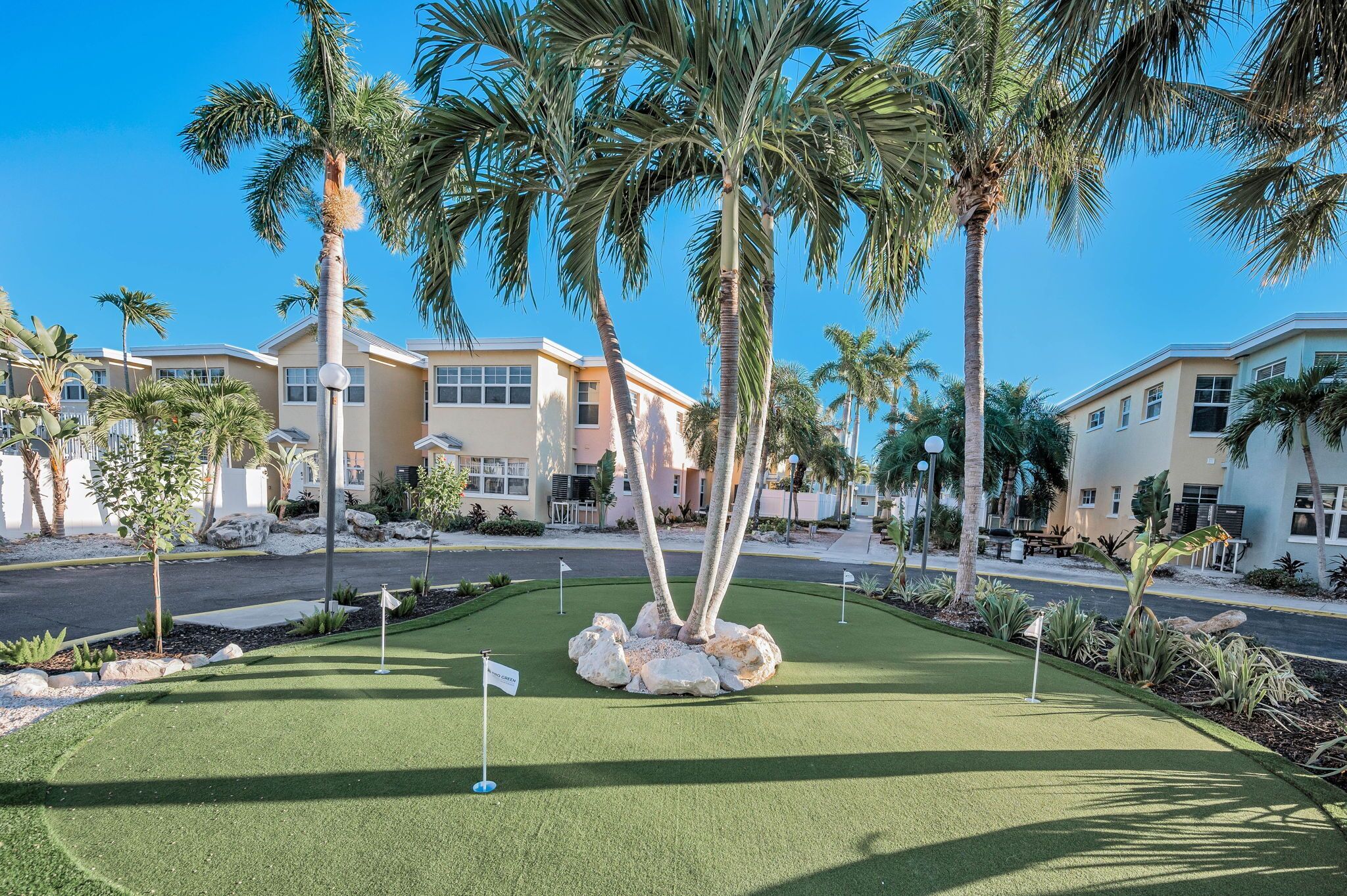 A well-maintained golf course with a putting green, surrounded by palm trees and modern buildings.