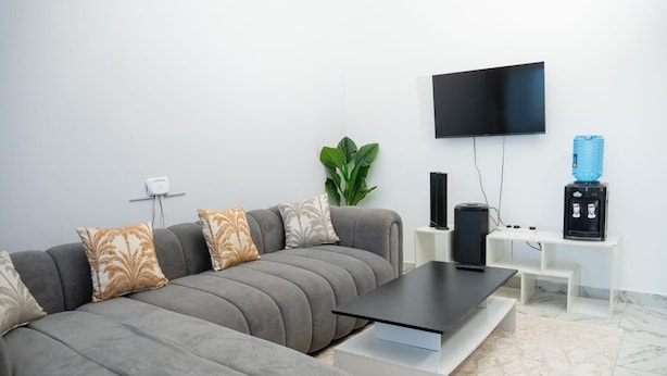 A modern living room with a grey sectional sofa, a flat-screen TV mounted on the wall, a water dispenser, and a speaker on a white console table. - ✨ Stylish Malindi 1BR Retreat | Cozy, Bright & Peaceful. Family -friendly (Malindi)