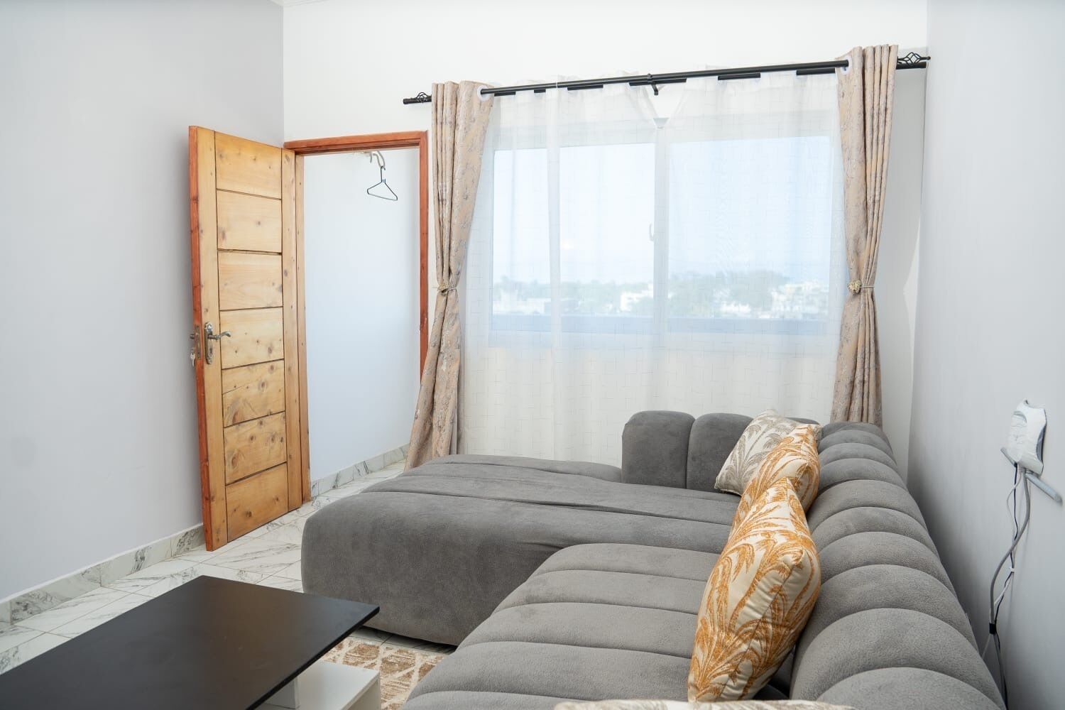 A room with a grey sofa, a wooden door, a window with sheer curtains, and a view of a cityscape.