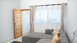 A room with a grey sofa, a wooden door, a window with sheer curtains, and a view of a cityscape. - ✨ Stylish Malindi 1BR Retreat | Cozy, Bright & Peaceful. Family -friendly (Malindi)