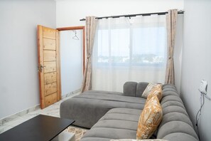 Una habitación con un sofá gris, una puerta de madera, una ventana con cortinas traslúcidas y vista a un paisaje urbano.