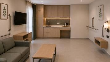 A modern hotel room with a sofa, a small table, a TV, a kitchenette, and a bathroom with grab bars.