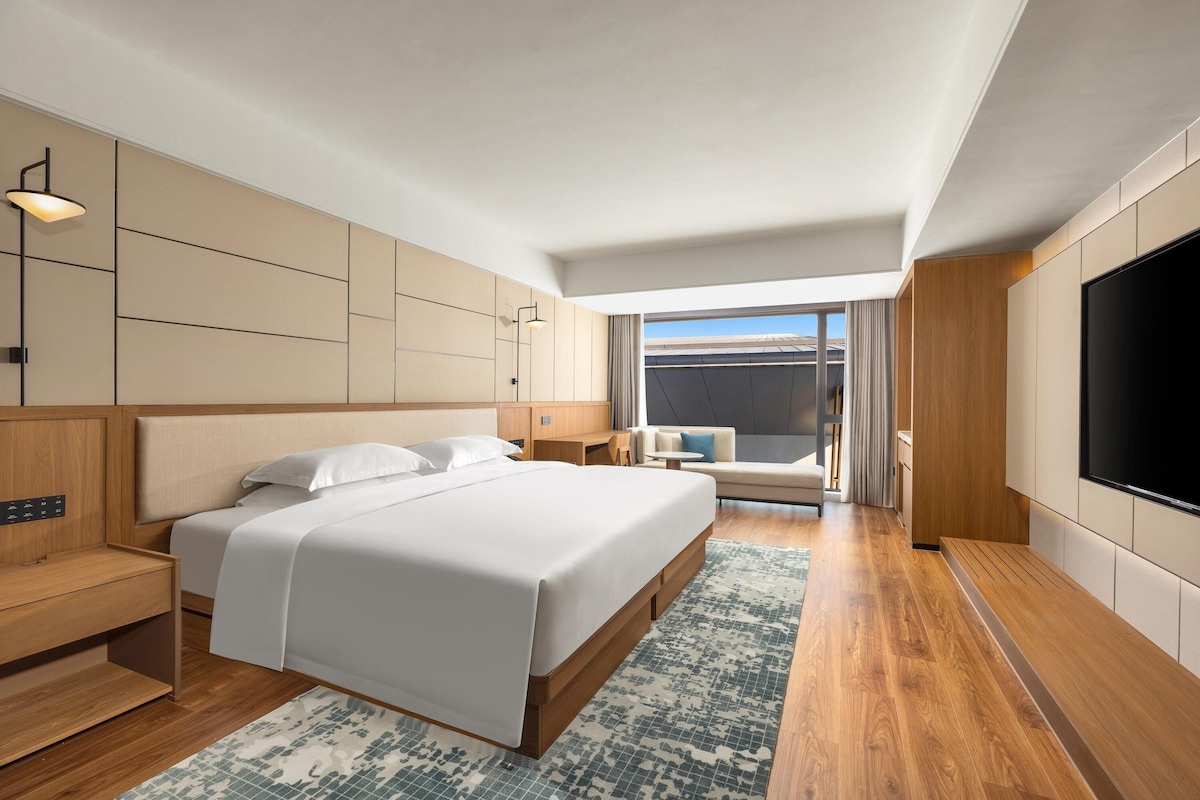 Quarto de hotel moderno com uma cama grande, televisão, área de estar e vista para o exterior.