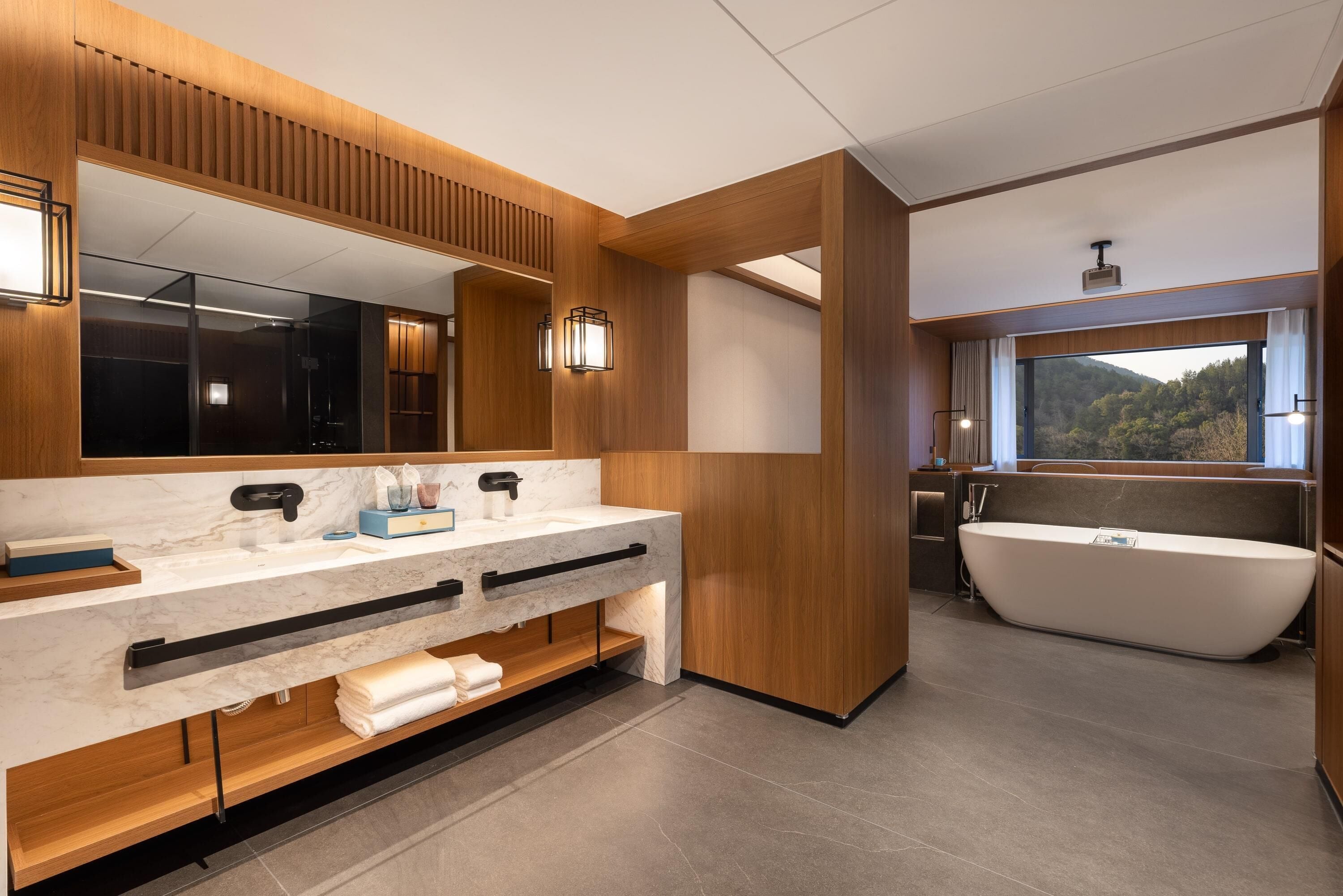 A modern bathroom with a large mirror, dual sinks, a freestanding bathtub, and a window with a view of the outdoors.