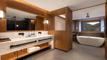 A modern bathroom with a large mirror, dual sinks, a freestanding bathtub, and a window with a view of the outdoors.