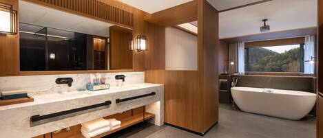 A modern bathroom with a large mirror, dual sinks, a freestanding bathtub, and a window with a view of the outdoors.