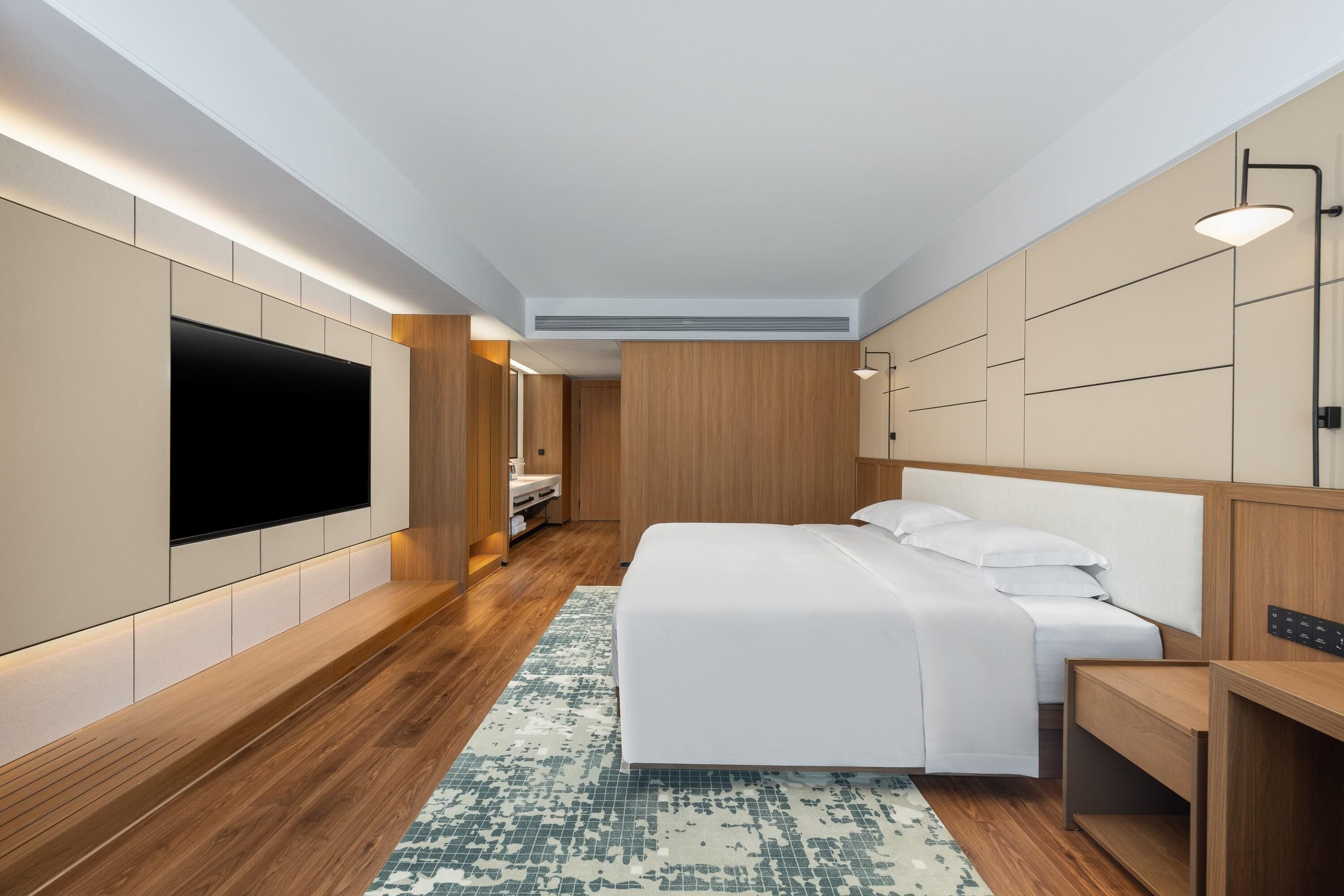 A modern hotel room with a large flat-screen TV mounted on the wall, a bed with white linens, a wooden headboard, and a small desk with a chair.