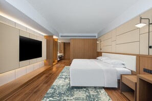 A modern hotel room with a large flat-screen TV mounted on the wall, a bed with white linens, a wooden headboard, and a small desk with a chair.