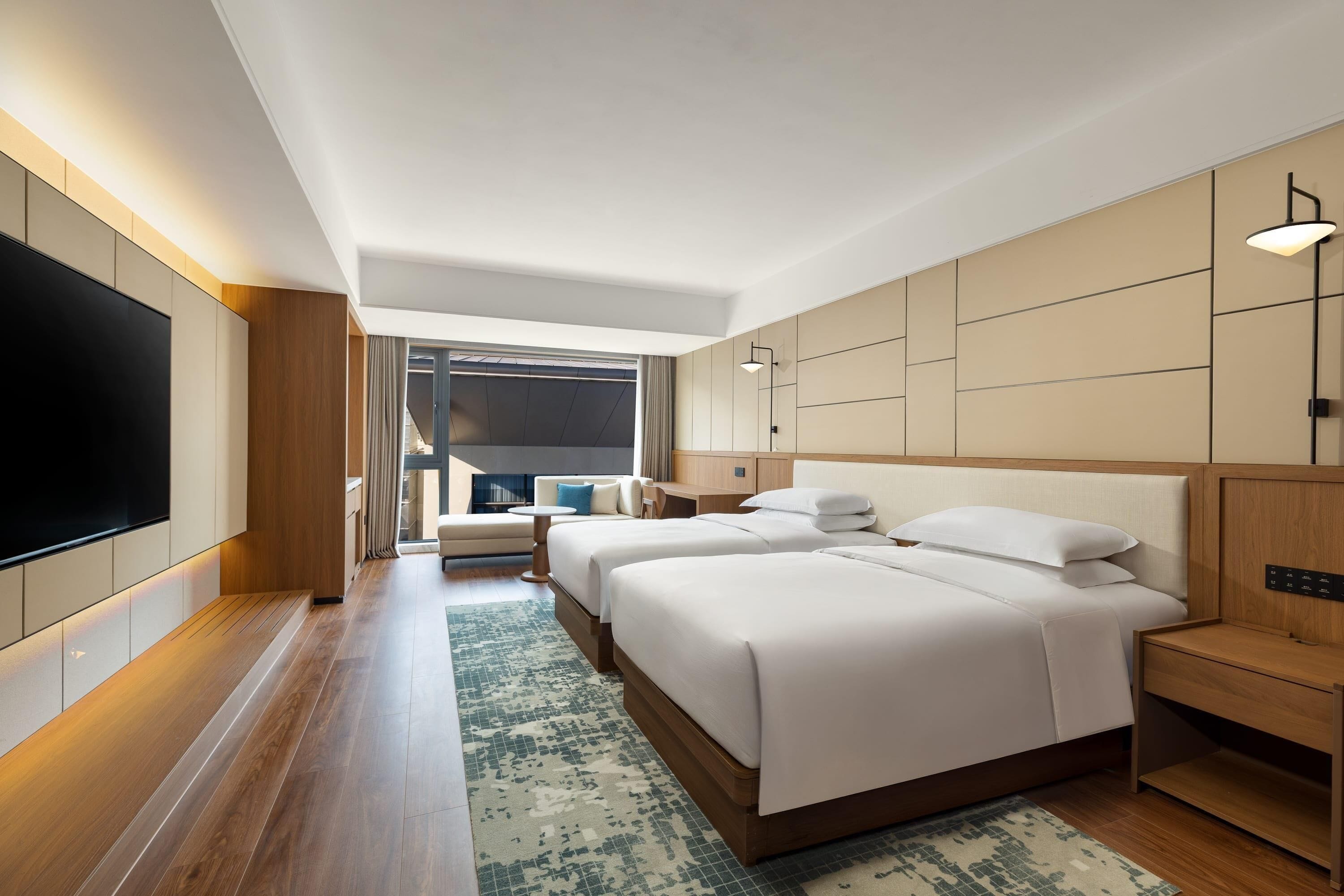 A modern hotel room with two beds, a flat-screen TV, and a large window.