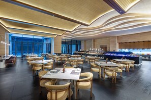 Uma sala de jantar ampla, com várias mesas e cadeiras, um buffet central e um design de teto moderno.