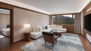 Habitación de hotel moderna con sofá, sillón y una mesa redonda de centro. Se aprecia una ventana grande con vistas a las colinas.
