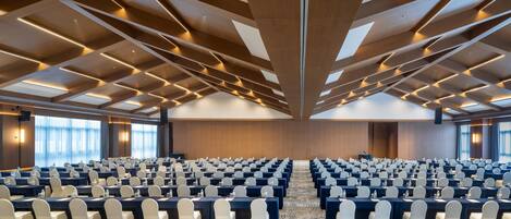 Un salón de conferencias con filas de mesas y sillas, una alfombra estampada y un techo alto con vigas de madera e iluminación.
