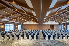 Un salón de conferencias con filas de mesas y sillas, una alfombra estampada y un techo alto con vigas de madera e iluminación.