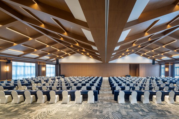 A conference room with rows of tables and chairs, a patterned carpet, and a high ceiling with wooden beams and lighting.