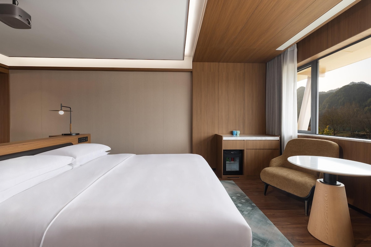 Quarto de hotel com cama, mesa de cabeceira, luminária, escrivaninha, cadeira e janela com vista.