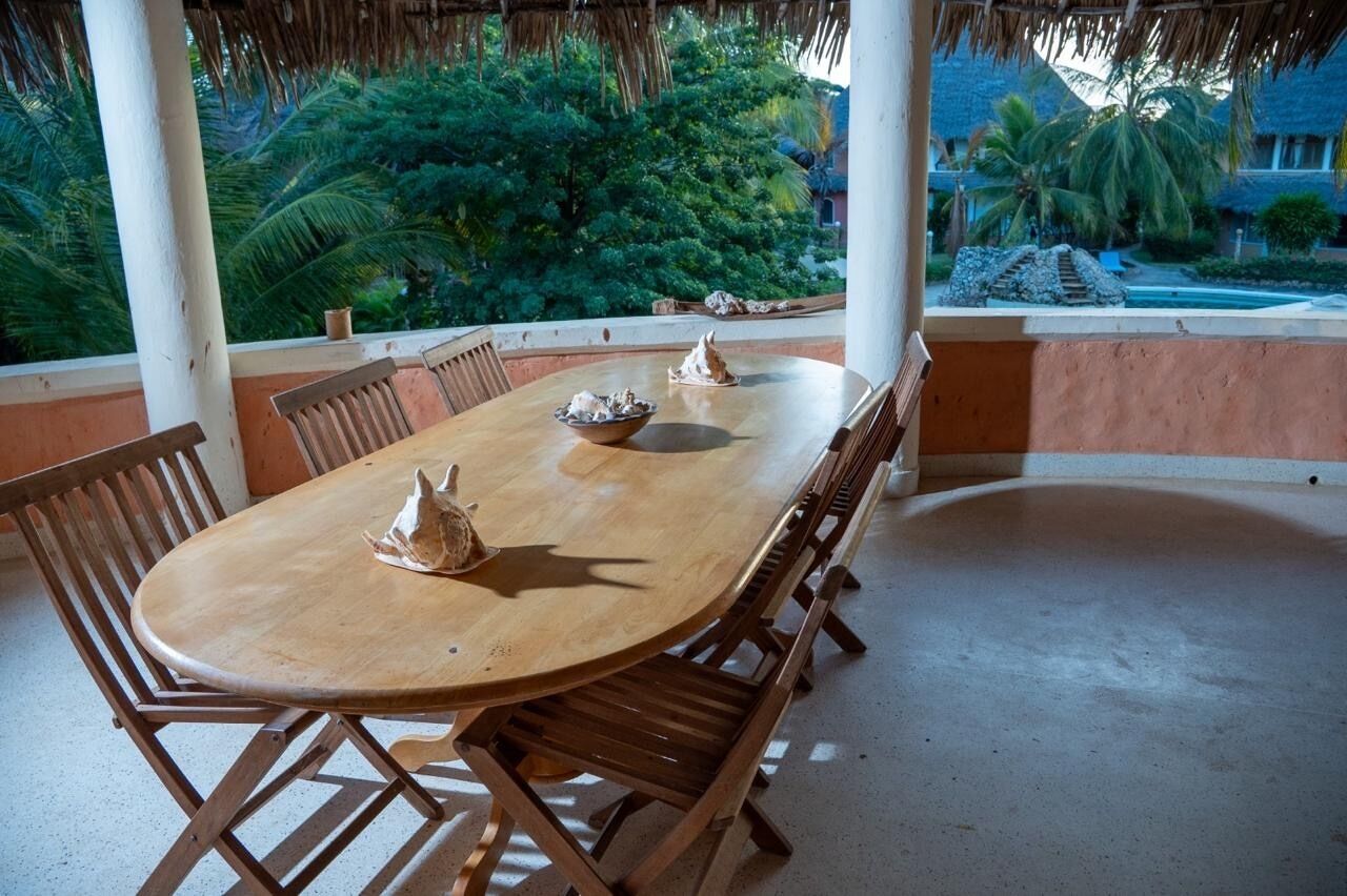 Une table à manger en bois avec des chaises, un toit de chaume et une vue sur une piscine et la verdure.