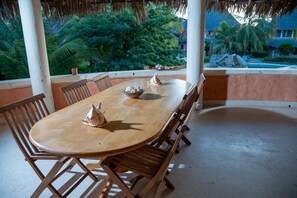 A wooden dining table with chairs, a thatched roof, and a view of a pool and greenery. - Tropical 2BR Malindi Condo | Pool & Balcony Views ❤️ (Malindi)