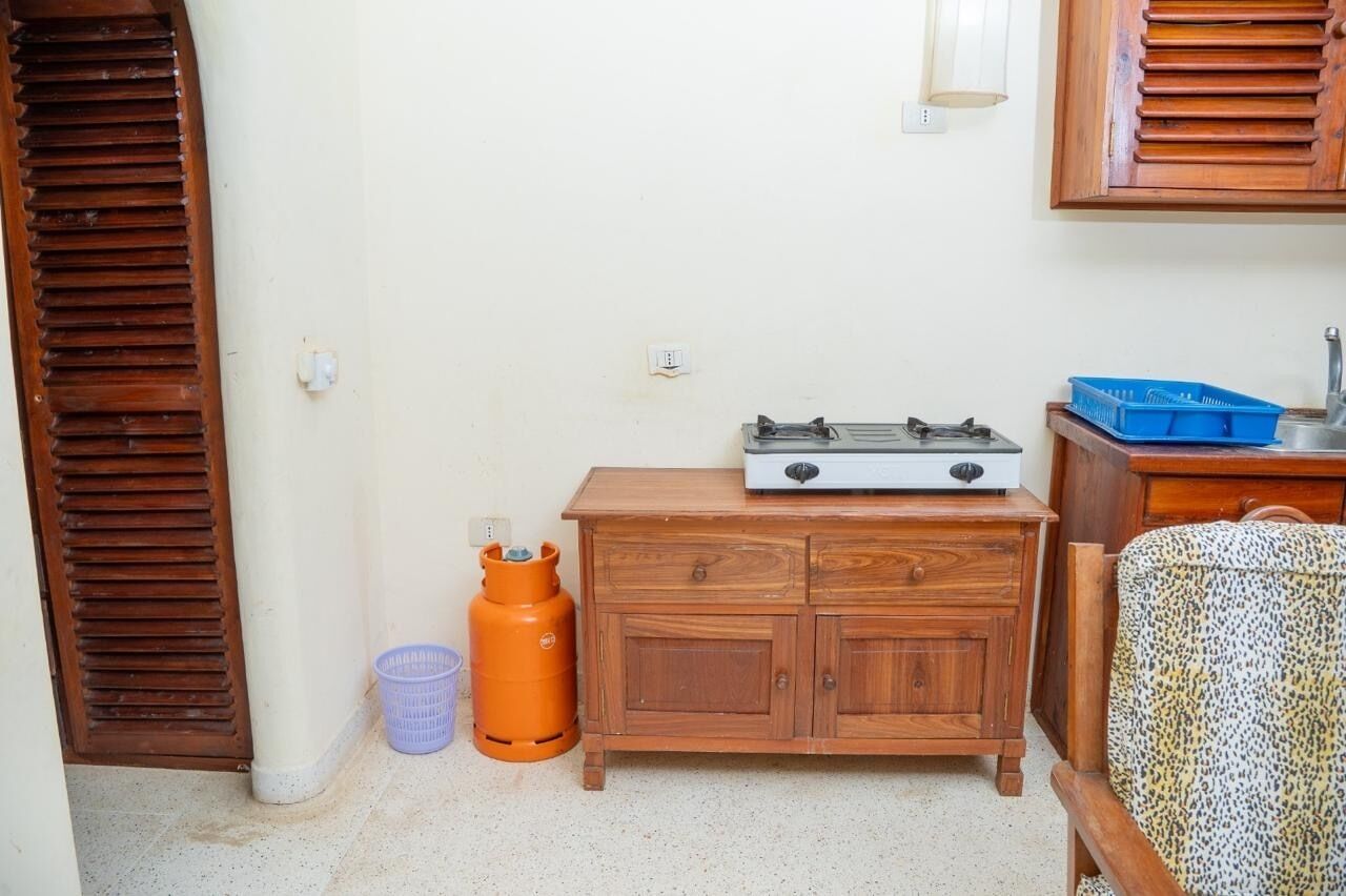 Une petite pièce comprenant un meuble en bois, une bouteille de gaz, une petite table avec une cuisinière à gaz et une chaise à motifs.
