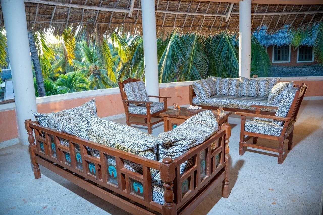 A covered outdoor seating area with a wooden sofa and chairs, a small table, and a view of palm trees.