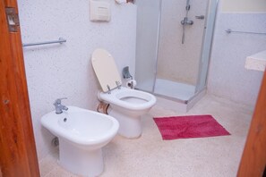 Casa de banho com duas sanitas, um bidé, um chuveiro e um tapete vermelho no chão.