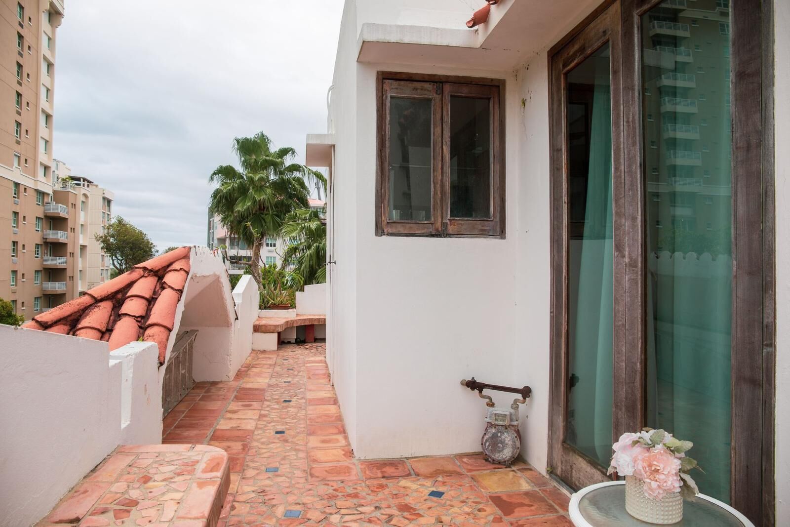 A narrow outdoor corridor with a tiled floor, a bench, a small table with a flower vase, and a door with a window.