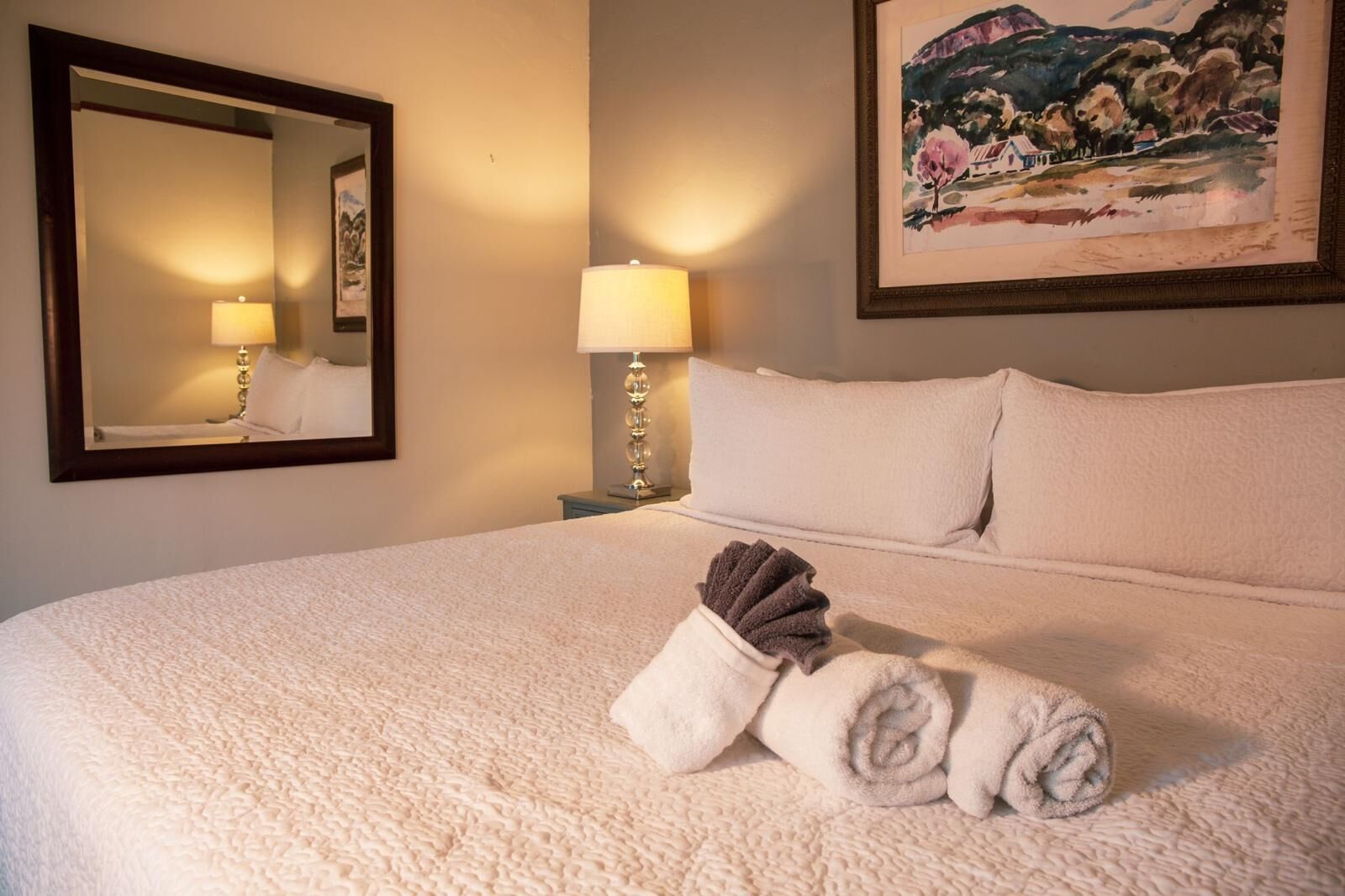 A hotel room with a large bed, a mirror, a painting, and a lamp.
