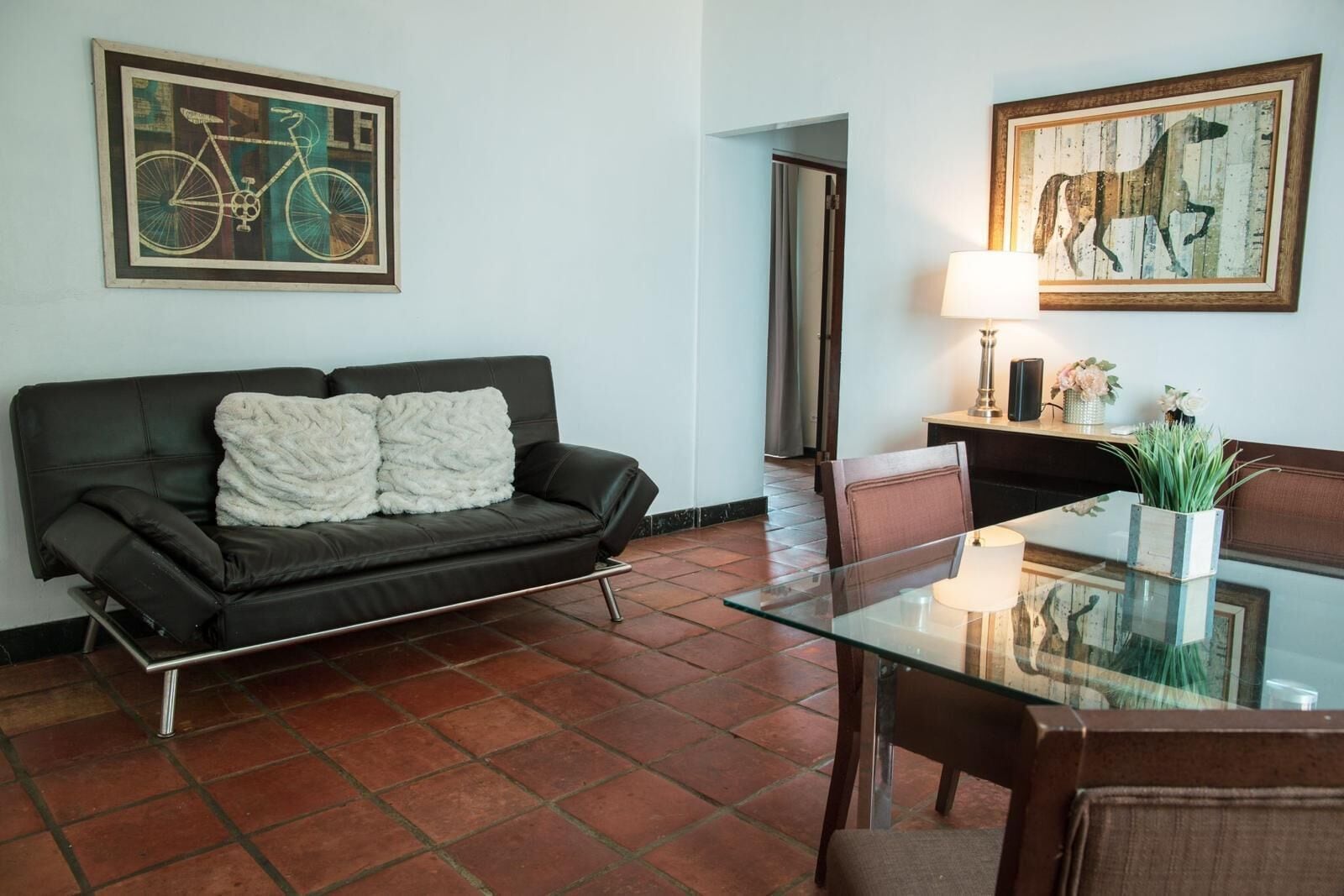A living room with a dark leather sofa, a glass coffee table, and a dining area with a chair and a lamp.