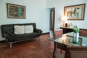 A living room with a dark leather sofa, a glass coffee table, and a dining area with a chair and a lamp. - Step to the beach /Beautiful Penthouse + Private Terrace (San Juan)