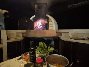 A kitchen with a wood-fired oven, a bowl of vegetables, a bottle of olive oil, and cooking utensils.