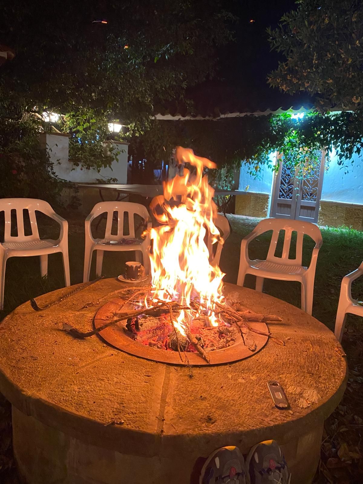 A round outdoor table with a fire pit in the center, surrounded by white plastic chairs.