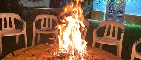 A round outdoor table with a fire pit in the center, surrounded by white plastic chairs.