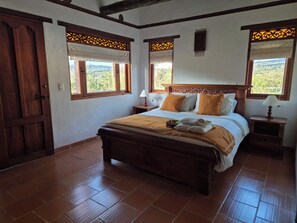 A bedroom with a wooden bed, two bedside tables with lamps, a window with a view of greenery, and a door.