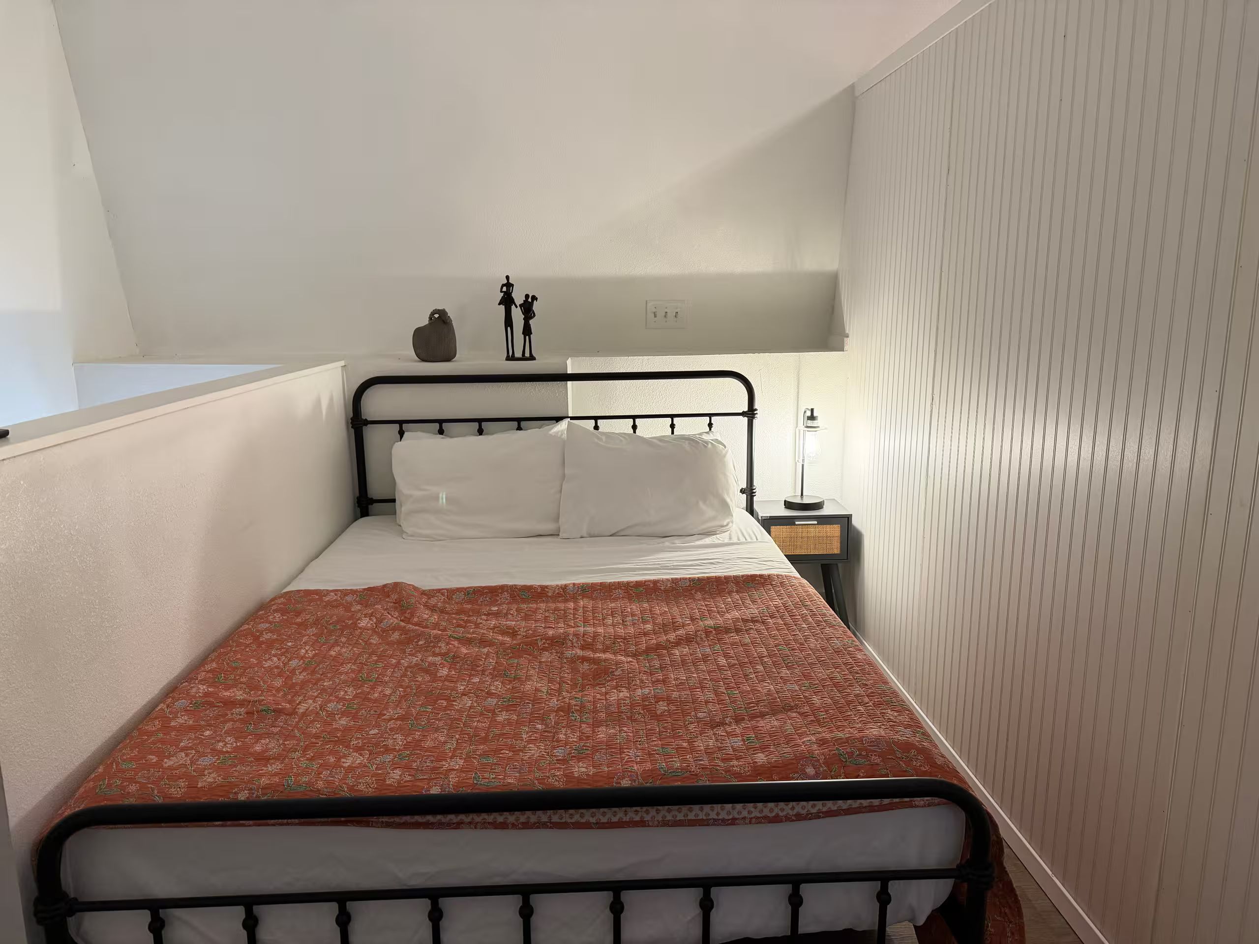 Een eenpersoonsbed met een bedekking met patroon, een nachtkastje met een lamp, en een wandplank met decoratieve objecten.