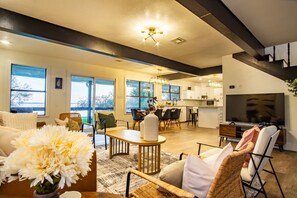 A spacious living area with a dining table, comfortable seating, a kitchen in the background, and large windows.