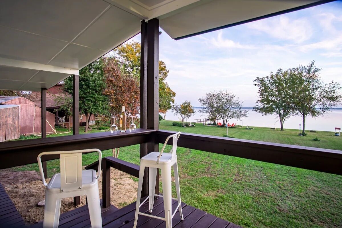 Un espace extérieur abrité comprenant deux chaises en métal blanc, une terrasse en bois et une vue sur une zone verdoyante avec des arbres et une étendue d’eau.