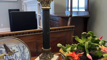 A lamp with a textured shade, a decorative plate with a bird and mountain scene, and a potted plant with green leaves and pink flowers on a wooden table.