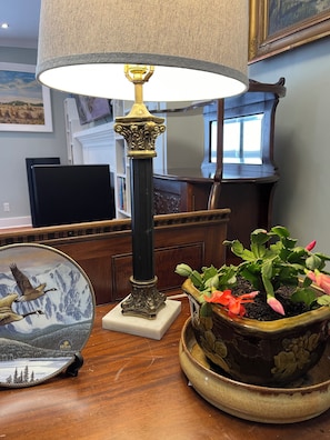 A lamp with a textured shade, a decorative plate with a bird and mountain scene, and a potted plant with green leaves and pink flowers on a wooden table.