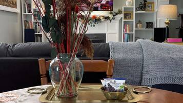 A living room with a sofa, a coffee table with a floral table runner, a vase with dried plants, and a television in the background.