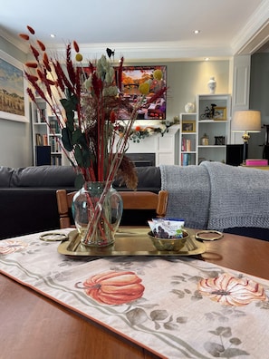 A living room with a sofa, a coffee table with a floral table runner, a vase with dried plants, and a television in the background.