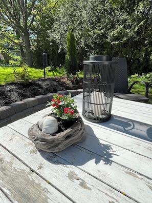 A wooden deck with a planter containing red flowers and white spherical stones, a metal trash can, and a bench.