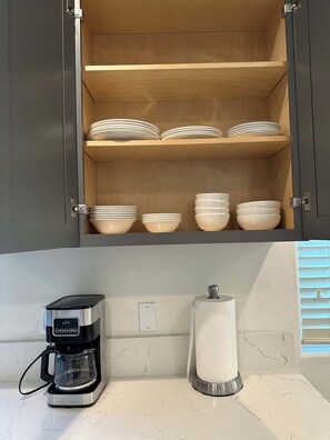 Una cocina con cafetera, dispensador de toallas de papel y un armario con tazones apilados.