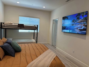 A bedroom with a bunk bed, a single bed, a large window with blinds, a flat-screen TV mounted on the wall, and a wooden floor. - 3BR Townhouse for Groups, Near Disneyland (Garden Grove)
