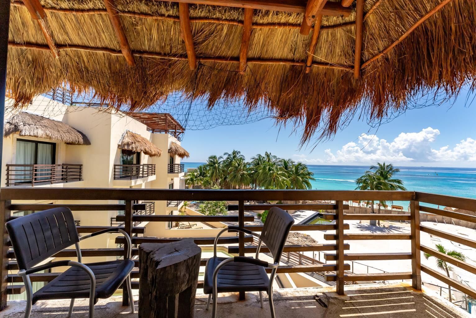 A balcony with a thatched roof overlooking a beach with palm trees and a clear blue sky.