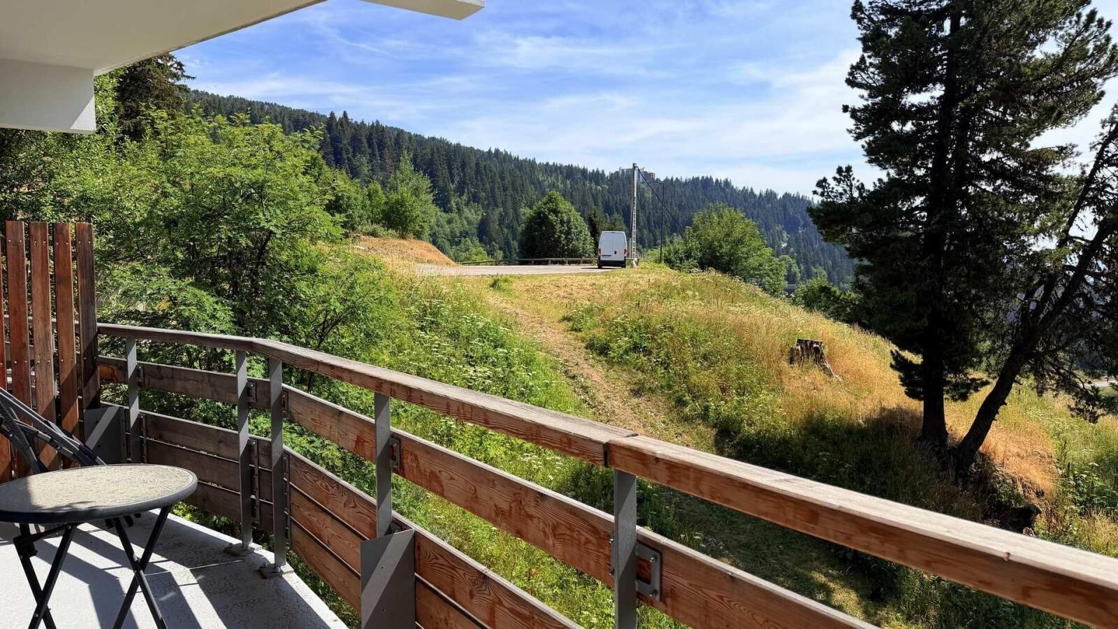 Un balcon avec vue sur une colline boisée et un fourgon blanc au loin.