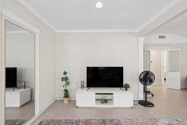 A modern living room with a flat-screen TV on a white entertainment unit, a standing fan, and potted plants.