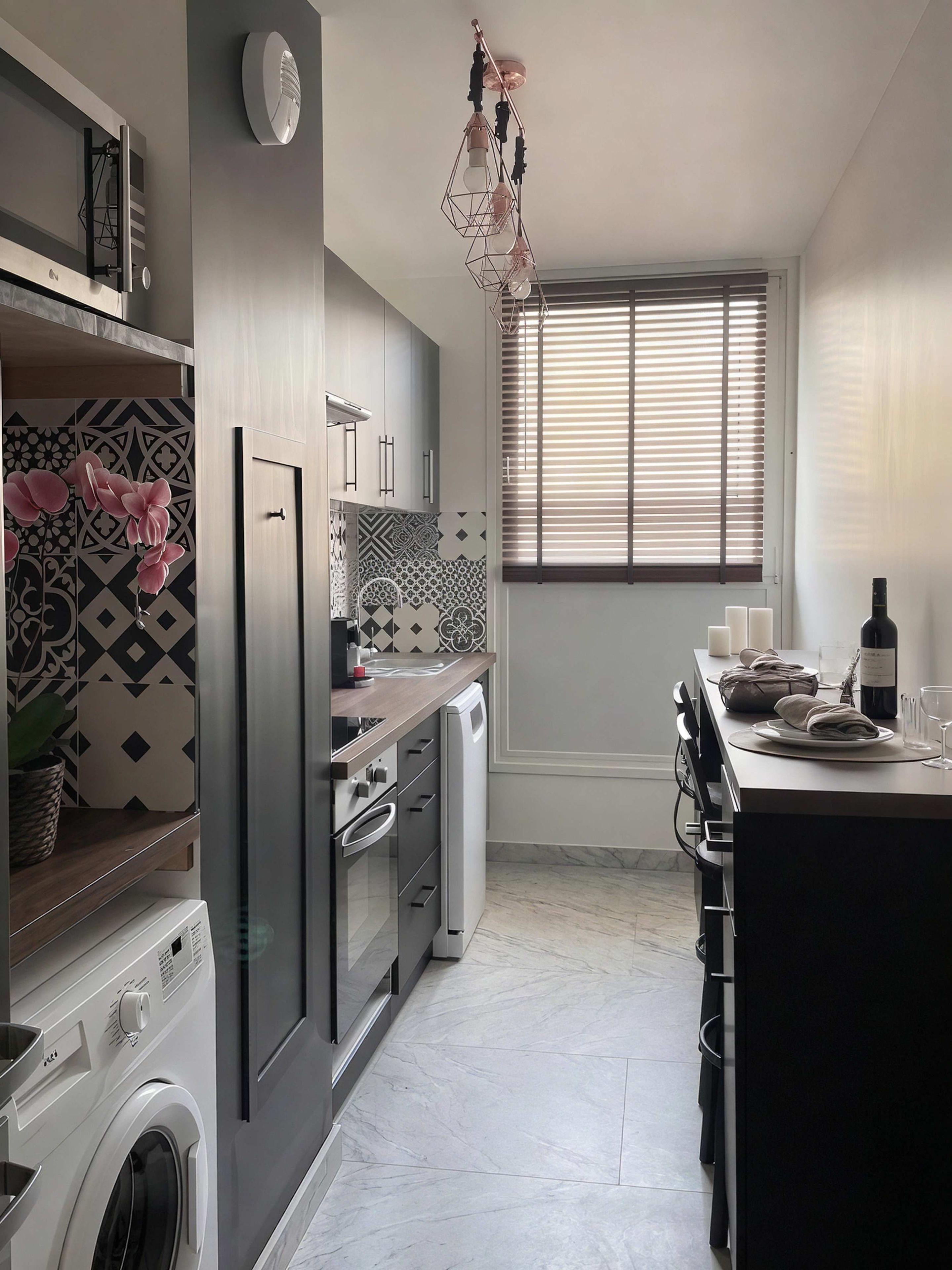 A modern kitchen with a built-in washing machine, a dining area with a table set for two, and a window with blinds.